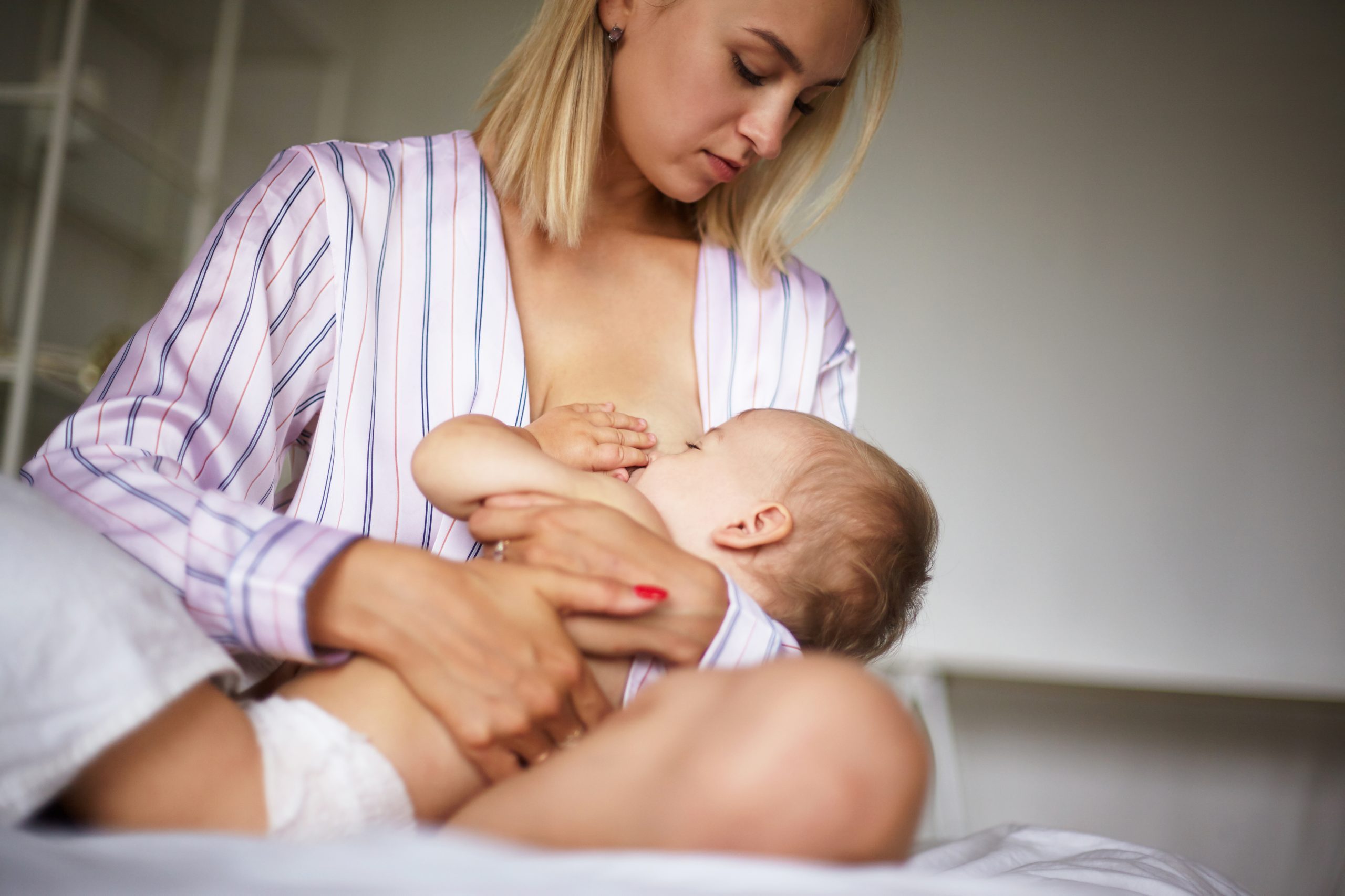 Tender beautiful young female wearing stylish silk pajamas sitting on bed embracing her baby, breastfeeding. Sleepy toddler sucking on mommy's breastmilk in bedroom. Childcare, love and care