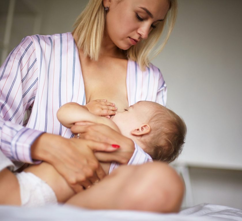 Tender beautiful young female wearing stylish silk pajamas sitting on bed embracing her baby, breastfeeding. Sleepy toddler sucking on mommy's breastmilk in bedroom. Childcare, love and care