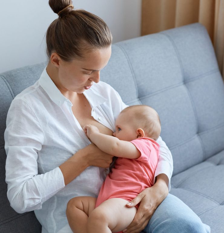 Portrait of young adult Caucasian woman with bun hairstyle wearing white shirt feeding her infant baby daughter, cute toddler baby eating and wants sleep.